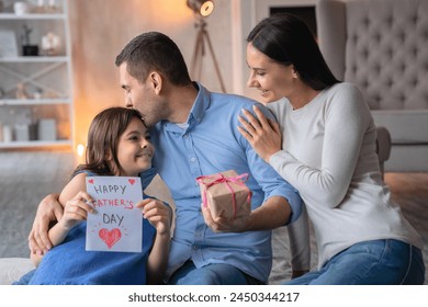 Happy father's day! Front view shot of happy family celebrating fathers day. Young man with a gift in hands his wife sitting on floor and hugging her husband. Little girl with present card for dad - Powered by Shutterstock