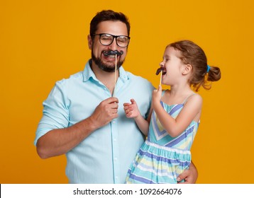 Happy Father's Day! Funny Dad And Daughter With Mustache Fooling Around On Colored Yellow Background
