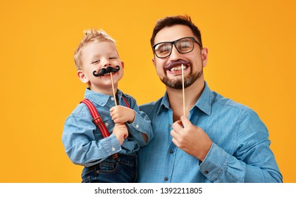 Happy Father's Day! Funny Dad And Son With Mustache Fooling Around On Colored Yellow Background
