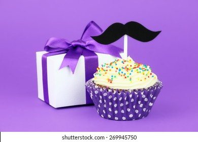 Happy Father's Day greeting with a gift box tied with purple ribbon and a cupcake decorated with buttercream frosting, colourful sprinkles and a black hipster mustache topper - Powered by Shutterstock