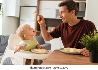 Happy Father's Day. Side View Of Dad Feeding Baby In Green Bib With Spaghetti During Lunch Time At Kitchen Table, Kid Sitting In High Chair Holding Fork. Happy Fatherhood, First Meal, Self-feeding