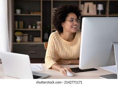 Happy female African business professional working at workstation monitor and laptop computer on desk, looking at screen, smiling, laughing. Young woman, employee, designer making video call