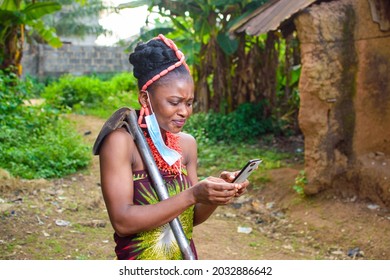 A Happy Female African Farmer Using Nose Mask And Also Have A Farming Hoe And Smart Phone With Her In A Village With Mud House And Banana Trees