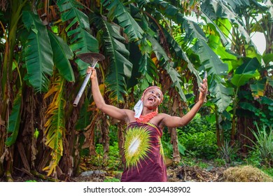 A Happy Female African Farmer Using Nose Mask, Having A Farming Hoe And Smart Phone With Her As She Spread Her Hands Wide On A Banana Farm