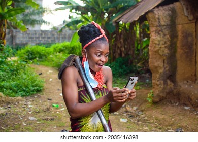 A Happy Female African Farmer Using Nose Mask And Also Have A Farming Hoe And Smart Phone With Her In A Village With Mud House And Banana Trees