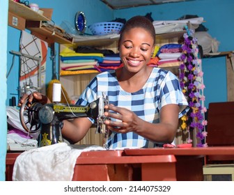 A Happy Female African Nigerian Tailor, Fashion Designer, Business Woman Or Entrepreneur Sitting Down With While Working With A Sewing Machine In A Tailoring Shop