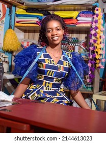 A Happy Female African Nigerian Tailor, Fashion Designer, Business Woman Or Entrepreneur Sitting Down With Tape Measure Around Her Neck In A Tailoring Shop