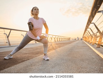 Happy female athlete in activewear enjoying workout in urban environment while doing stretching exercise. Sportswoman stretching legs on a treadmill on a city bridge while working out outdoor - Shutterstock ID 2114355581