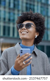 Happy Female Blogger Uses Cellphone For Finding Route Strolls In City Wears Sunglasses And Grey Jacket Focused Above With Glad Expression Installs Application For Tracking Location Has Travel Vacation