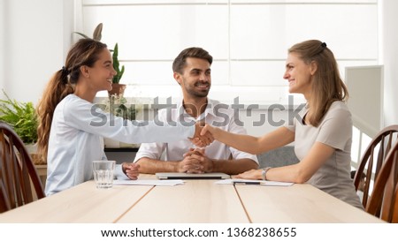 Happy female business partners shake hands sign two contracts with lawyer mediation at meeting, smiling businesswomen handshake make legal partnership deal after successful negotiations with mediator