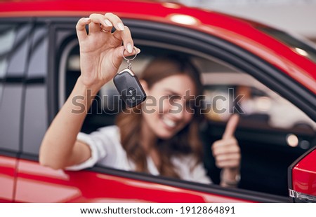Happy female client smiling and gesturing thumb up while sitting in new vehicle and demonstrating keys in showroom