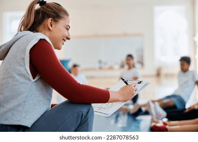 Happy female coach analyzing exercise plan while having physical education class with group of kids at school gym. - Powered by Shutterstock