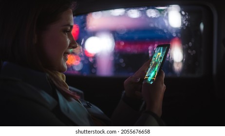 Happy Female Is Commuting Home In A Backseat Of A Taxi At Night. Beautiful Woman Passenger Playing Arcade Video Game On Smartphone While In A Car In Urban City Street With Working Neon Signs.