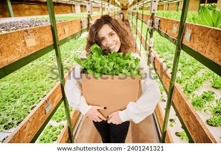 Happy female customer looking at camera and smiling while holding cardboard package box with green leafy plants. Woman with fresh leafy greens standing near shelves with plants in greenhouse.