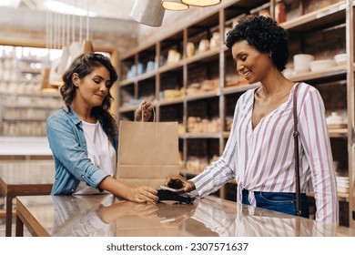 Happy female customer making a contactless NFC payment using her smartphone. Satisfied young woman smiling cheerfully while shopping from a local female-owned ceramic store. - Powered by Shutterstock