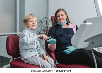 Happy Female Dentist Sitting In Dental Chair And Showing To Patient How Not To Fear Working Drill, Young Boy Holding The Tool And Smiling, Healthy Teeth Concept