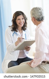 Happy Female Doctor With Clipboard Looking At Patient In Clinic