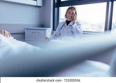 Happy Female Doctor Sitting In Hospital Room And Making A Phone Call. Medical Professional In Hospital Ward Using Mobile Phone.