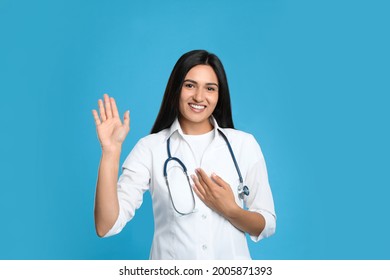 Happy Female Doctor Waving To Say Hello On Light Blue Background