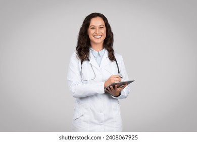 Happy female doctor in white lab coat using tablet, showcasing modern medical technology or patient records access, grey background - Powered by Shutterstock