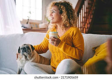 Happy female dog owner smile and enjoy happy time at home with her best friend old pug sitting near her. Animal and love therapy with people. Adult woman have care of her puppy and drink a tea - Shutterstock ID 2171905659