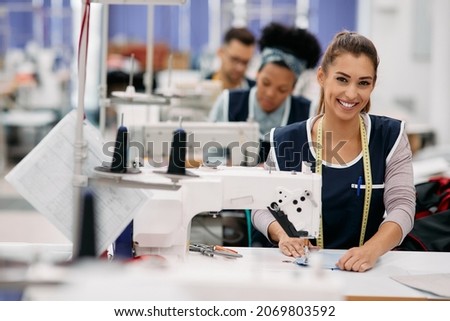 Happy female dressmaker working with sewing machine at textile factory and looking at camera. 