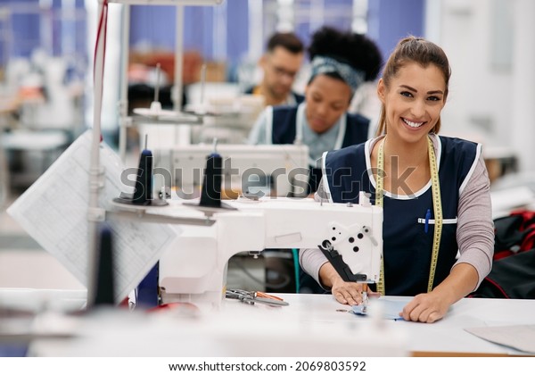 Happy female dressmaker working\
with sewing machine at textile factory and looking at camera.\

