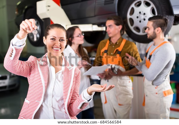 Happy female driver has just got keys from her\
repaired car at a car repair shop. Service crew is standing near\
lifted up car in\
background