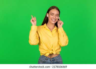 Happy Female In Earbuds Listening To Music Posing Standing With Eyes Closed Over Green Studio Background. Woman Enjoying Favorite Song Wearing Earphones. Great Playlist Concept