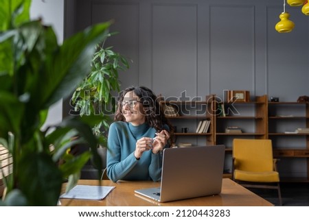 Happy female employee looking in window with satisfied face expression while sitting in office in front of laptop, woman taking break from computer work at workplace. Job satisfaction concept
