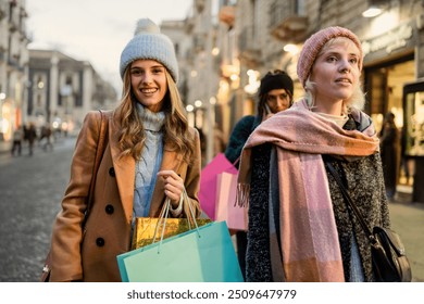 Happy female friends enjoying winter shopping in city center. Stylish women wearing warm coats and hats, carrying colorful bags on illuminated street. Urban lifestyle and friendship concept. - Powered by Shutterstock