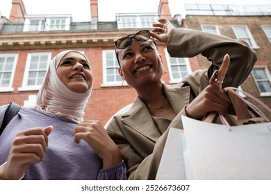 Happy female friends in front of window shop and pointing items. They are a multicultural couple, one is muslim wearing hijab and the other is mixed race with short hair and some shopping bags. - Powered by Shutterstock