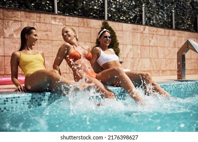 Happy Female Friends Splashing Water While Sitting At The Egde Of A Swimming Pool.