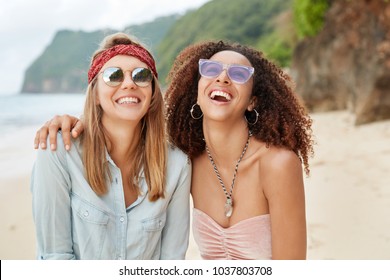 Happy Female Gay Couple Of Different Nationalities, Embrace Each Other, Laugh Joyfully And Pose At Camera Against Cliff And Seascape. Same Sex Family Has Summer Resort Together. People And Relations