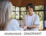 Happy female holistic teacher holding hands of woman during spiritual practice meditation experience feeling energy, peace and love, giving support during body health retreat healing training session.