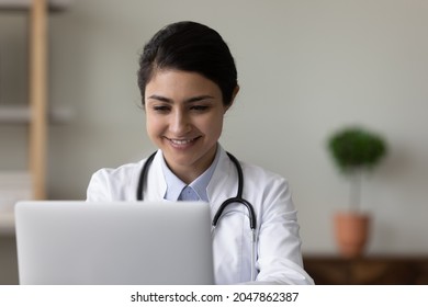 Happy female Indian doctor chatting online with patient, giving virtual consultation, making video call on laptop computer. Young therapist working at computer in clinic office, smiling at screen