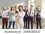 Happy female leader receives business award. Smiling woman winner holding golden trophy, with team of joyful employees in background cheering, celebrating and congratulating her on great work success