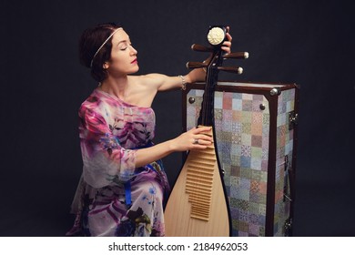 A Happy Female Musician In Asian Clothes On A Studio Black Background. A Smiling Woman With A Stringed Musical Instrument From Asia On A Dark Background