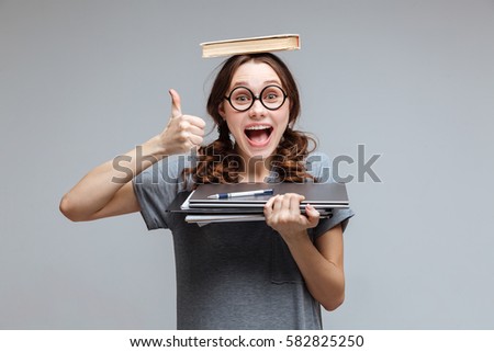 Happy Female nerd in funny eyeglasses with book on head which holding laptop and notebook in hand and showing thumb up