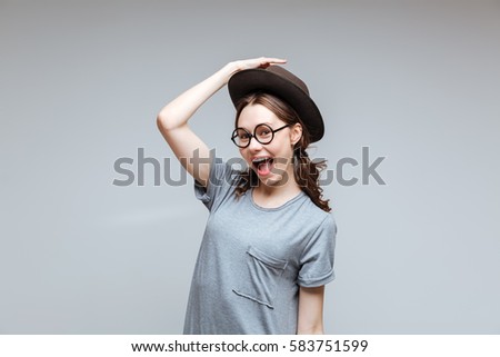 Happy Female nerd in glasses which holding for her hat and looking at camera. Isolated gray background