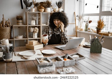 Happy female with new purchase in ceramics shop sitting at table against background of shelves with dishes. Young curly woman potter taking order for purchase of tableware using phone and laptop - Powered by Shutterstock