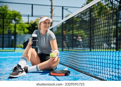 Happy female paddle tennis player drinking water after playing a match on outdoor court. - Powered by Shutterstock