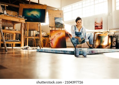 Happy female painter making a blue painting on a canvas. Creative young woman smiling while looking at her painting on the floor. Cheerful young artist working on a new project in her atelier. - Powered by Shutterstock