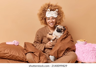 Happy female pet owner with cute pug dog wrapped in blanket wears pajama and sleepmask plays with favorite pet pose in bedroom against brown background. Domestic atmosphere animals friendship