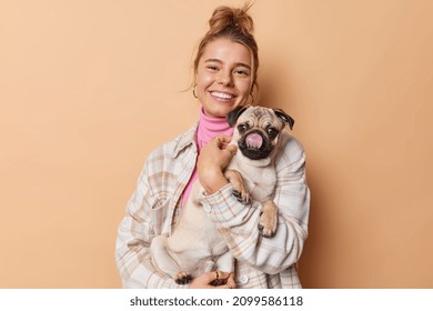 Happy Female Pet Owner Dressed Casually Holds Pug Dog Which Sticks Tongue Spends Free Time With Favorite Puppy Isolated Over Beige Studio Background. People Animals And Relationship Concept.