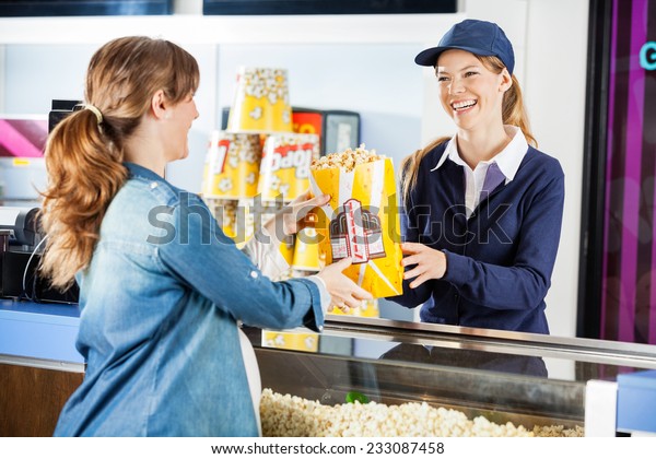 Happy female seller giving popcorn\
paperbag to pregnant woman at cinema concession\
stand