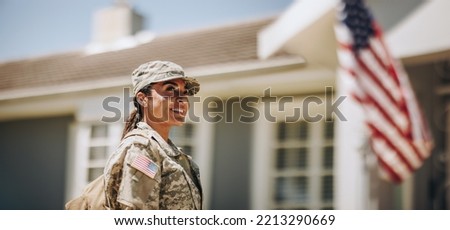 Happy female soldier looking away with a smile while standing outside her house with her bag. American servicewoman coming back home after serving her country in the military.