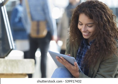 A happy female student smiles as she uses her digital tablet outdoors. Her friends are in the background out of focus.