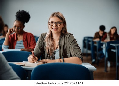 Happy female student taking notes while attending a lecture at the university and looking at camera. - Powered by Shutterstock