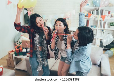 Happy Female Students Enjoy House Party Celebrating Graduation Playing With Blowers. Carefree Girls Dancing Raising Hands With Toys And Music In The Cozy Living Room In Decorated Home.